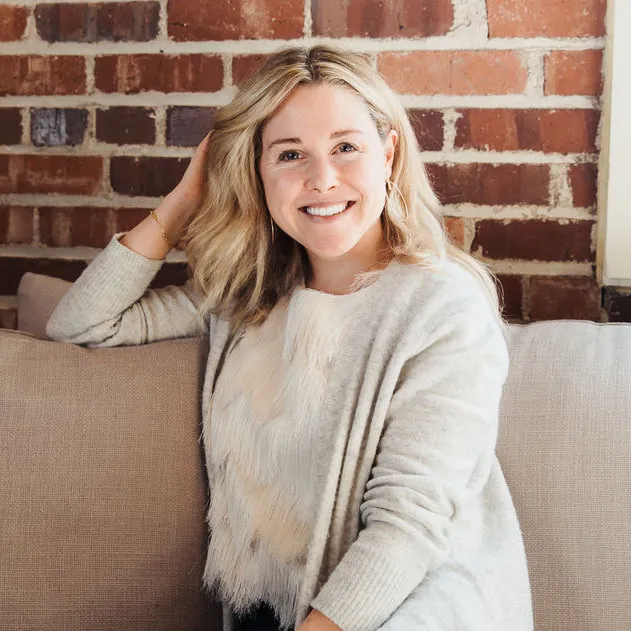 Smiling woman with blonde hair sitting on a beige couch, wearing a light gray cardigan and white top, with a brick wall background.
